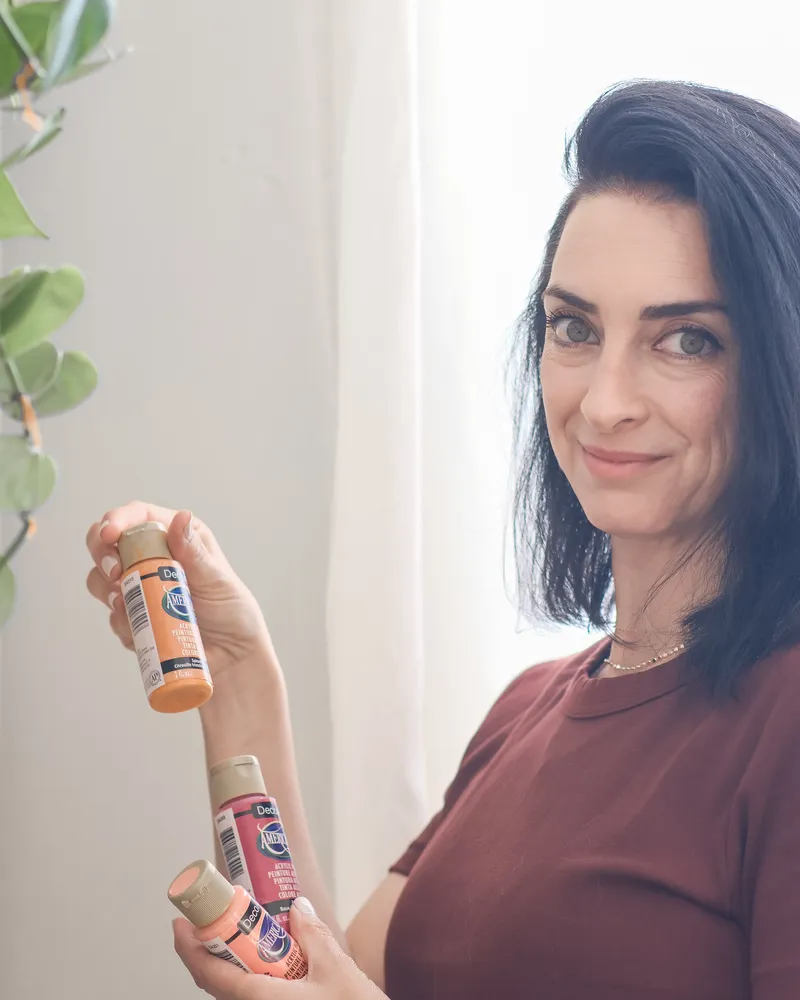 אלה ברקת בן ארי מחייכת ומחזיקה בקבוקוני צבע אקרילי
