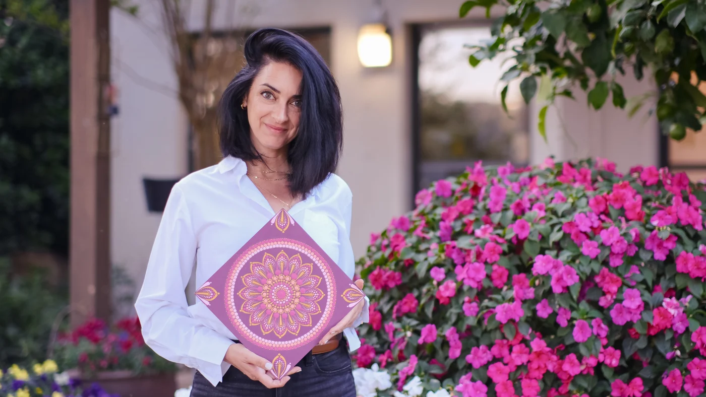 אלה ברקת בן ארי מחזיקה מנדלה מנוקדת בגווני ורוד וזהב ליד שיח פרחים ורודים